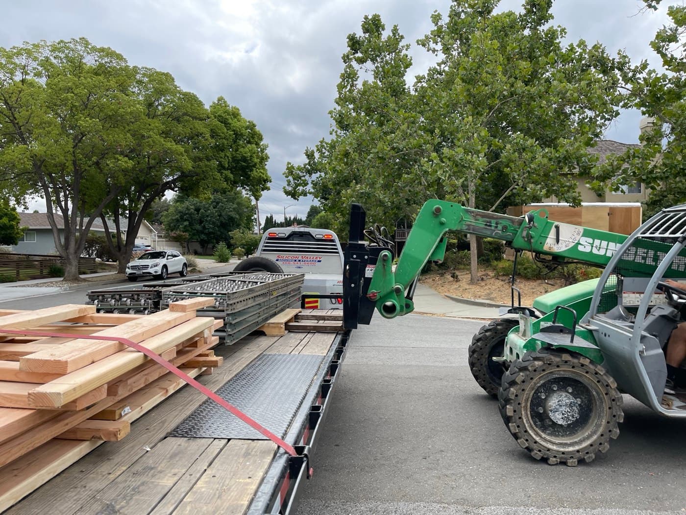 Telehandler with pallet forks raised beside a loaded SVM flatbed trailer on a tree-lined street