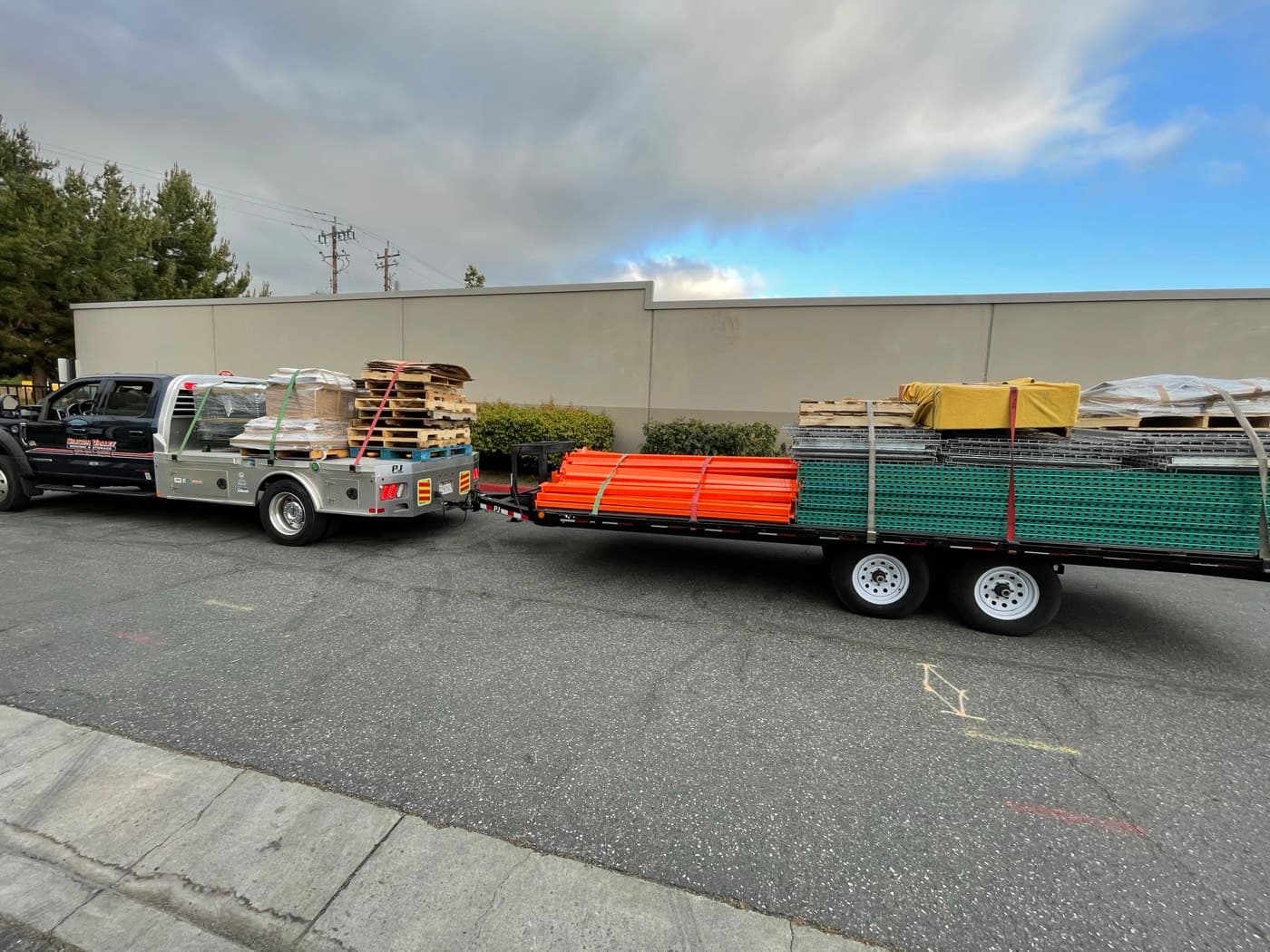SVM flatbed truck and trailer loaded with orange pallet rack beams, green uprights, wire decking, and pallets, strapped down outside a warehouse